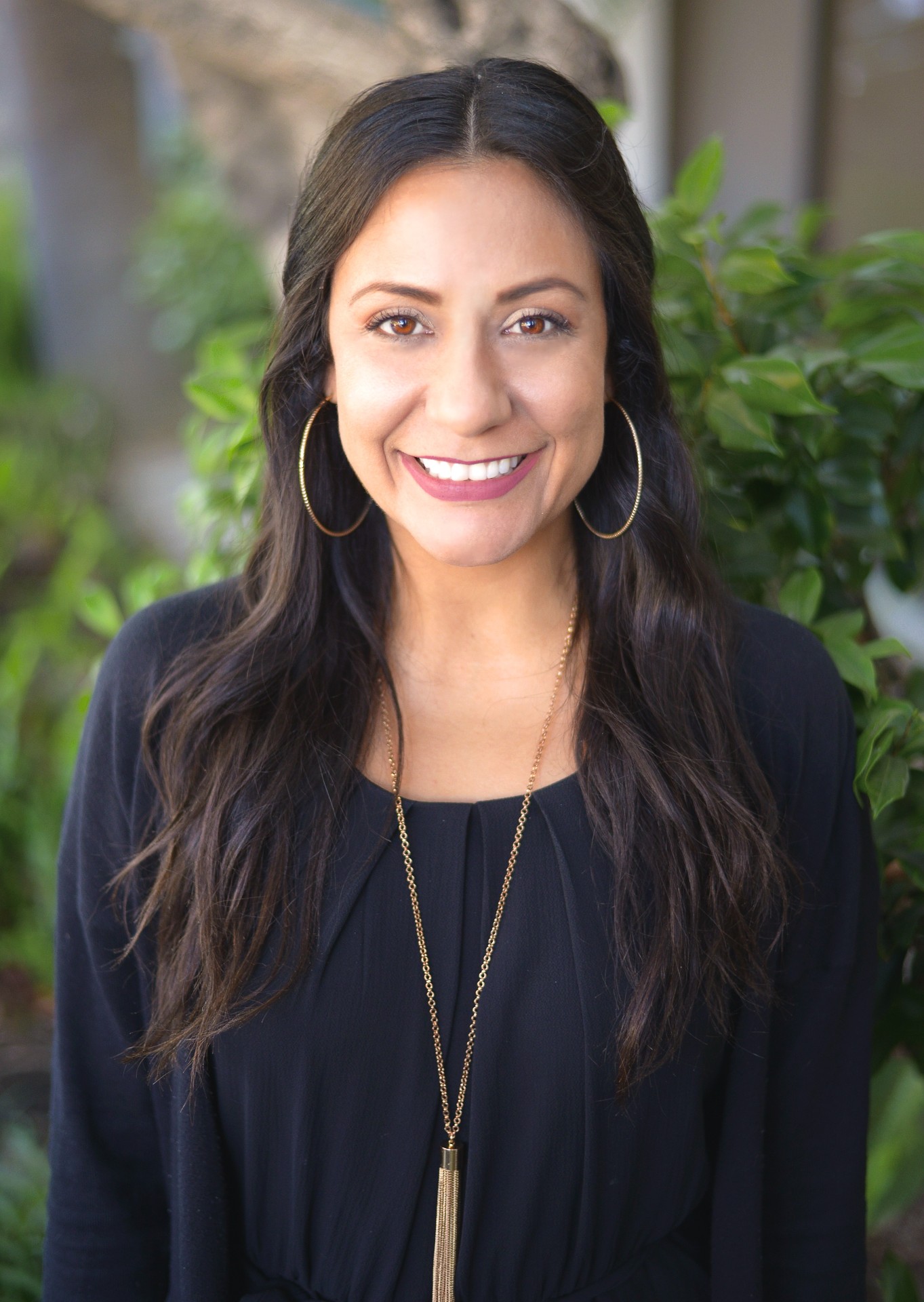 Portrait of Dee Rossiter who is smiling and wearing a black long-sleeve top with a long golden necklace, in front of a green plant