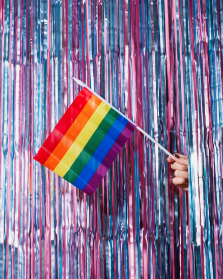 A hand holding a LGBTQ+ Pride flag pokes through blue and pink streamers.