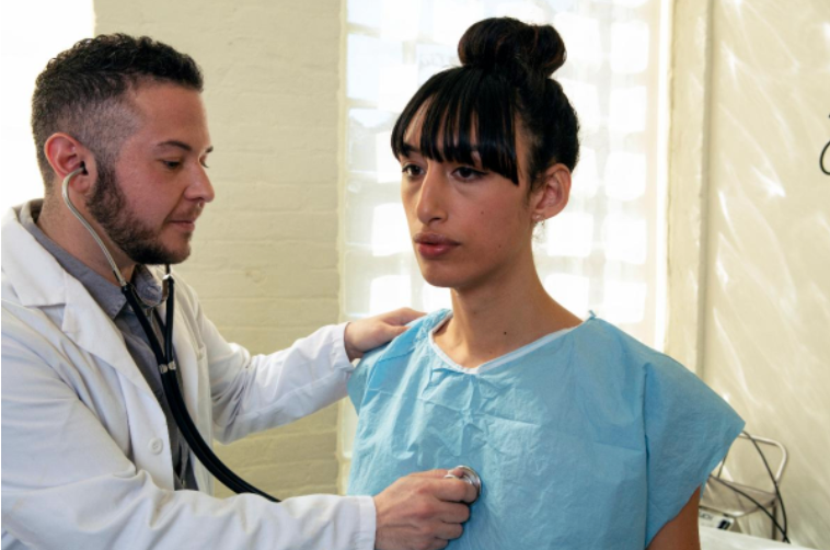 A transgender woman in a blue hospital gown being treated by a doctor, a transgender man