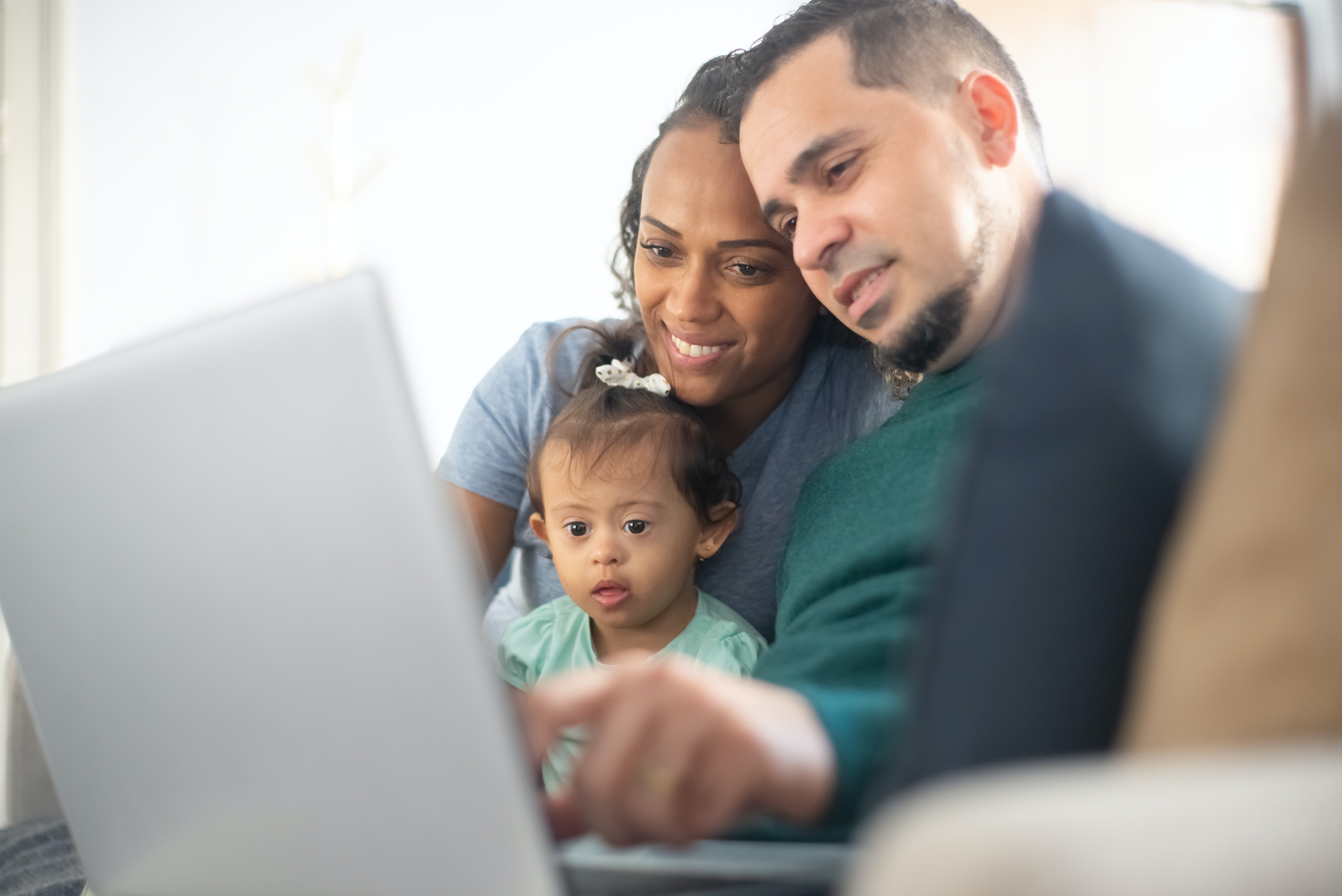 Two parents sit together in front of a laptop. Their baby sits on the mother's lap and stares at the screen while the father points at the screen.