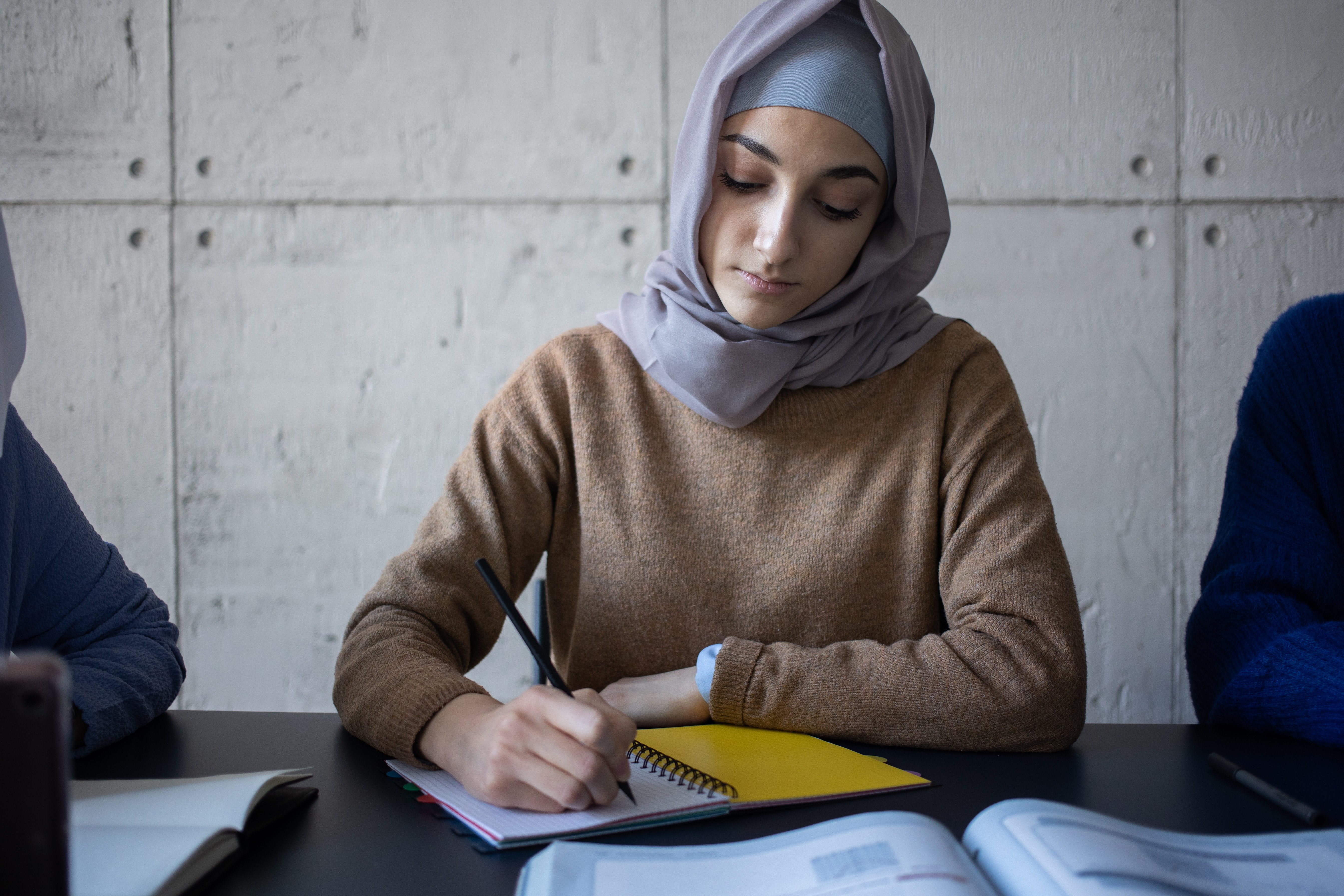 A person with a beige sweater and light purple headscarf takes notes in a notebook.