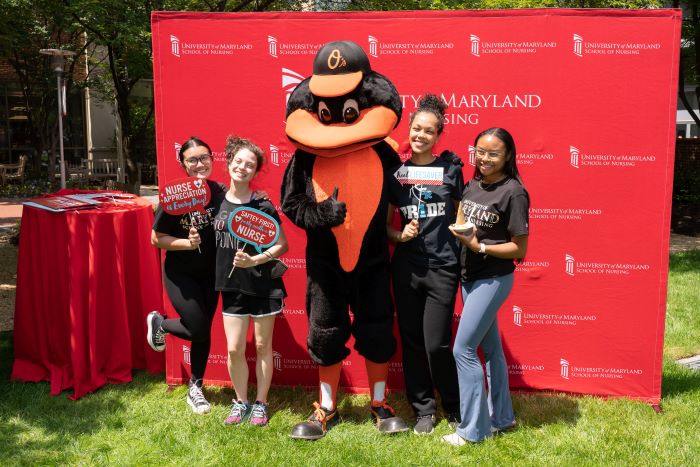 students with Oriole Bird holding photo booth props in front of UMSON step and repeat