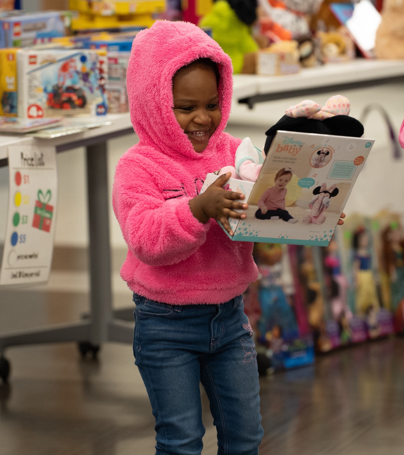 Little girl in pink sweater with hood up holding minnie mouse doll and smiling at the Christmas Store