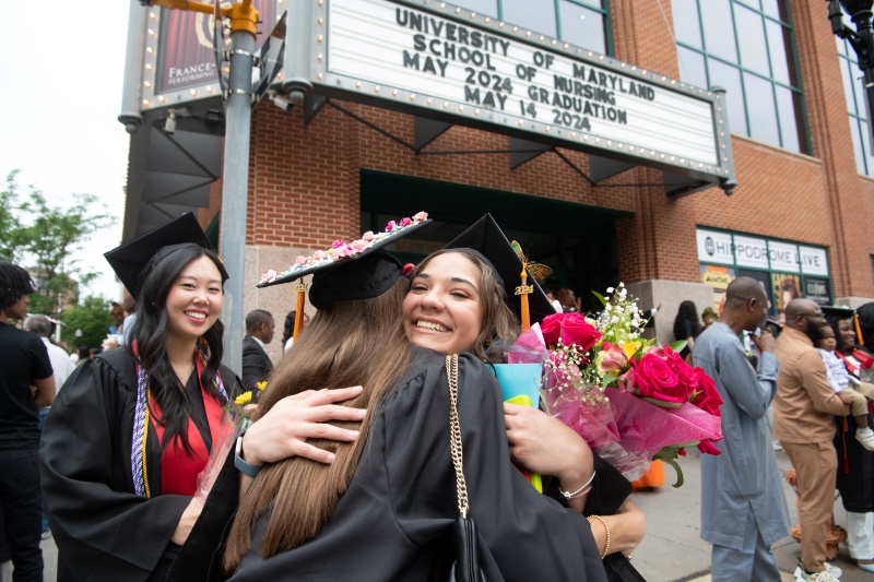 image of two graduates hugging, one holding flowers, under marquee at Hippodrome Theatre announcing UMSON graduation