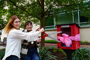 Sarah Michel and Ariana Campos holding giant scissors in a ribbon cutting of the Little Free Library.