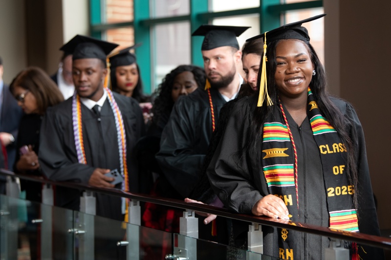 graduates in regalia lined up at bannister; first graduate is smiling at camera, wearing stole that says 