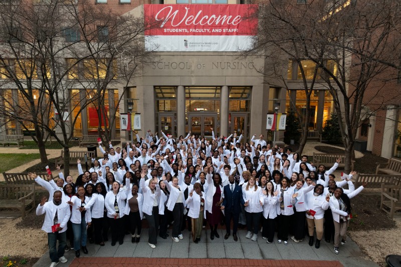 large group of entry-into-nursing students and faculty wearing white scrub jackets, throwing their hands up in the air, in front of the School of Nursing Building, with Welcome banner hanging behind
