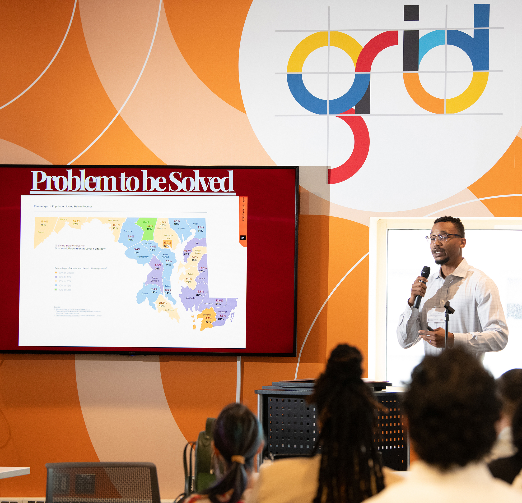 man holding microphone and speaking in front of the grid sign