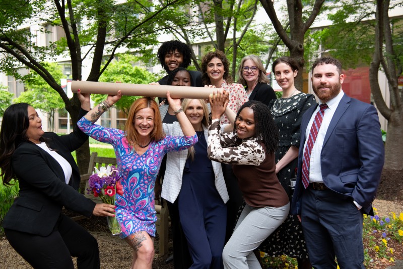 A diverse group of smiling people in formal attire joyfully hold a large cardboard tube outdoors, surrounded by lush greenery, conveying celebration.