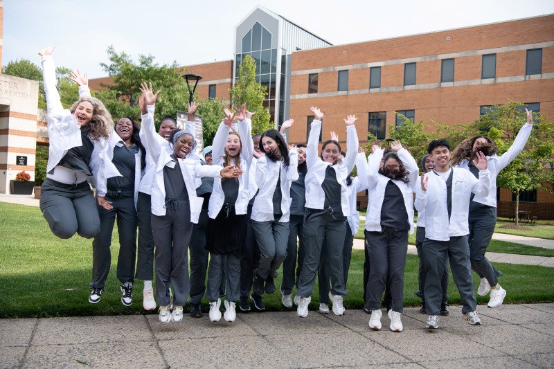 Joyful group of diverse students in white scrub jackets and gray uniforms jump with arms raised outside a brick school building.