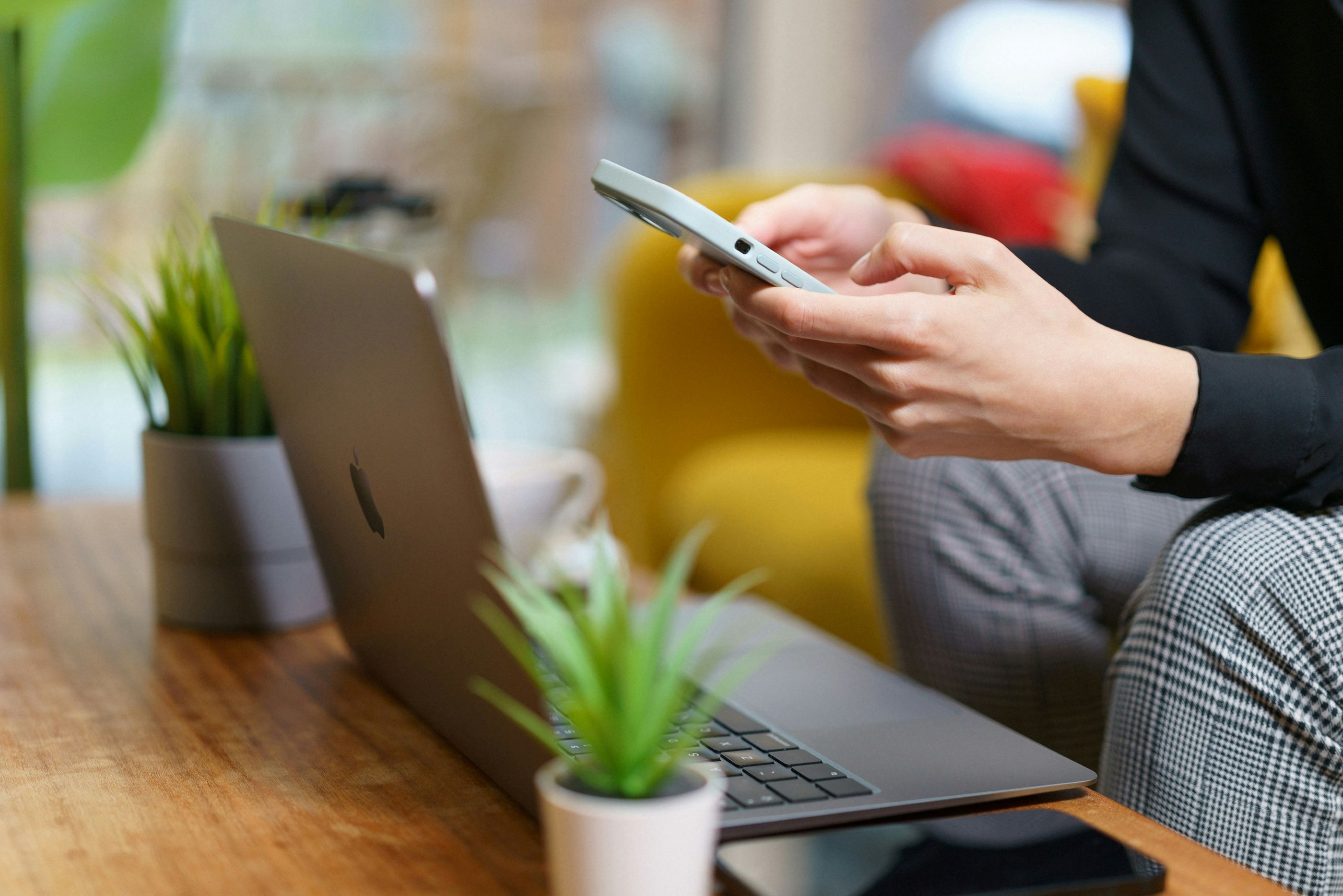 A person sitting on a sofa types on a cell phone with a laptop placed on a coffee table in front of them