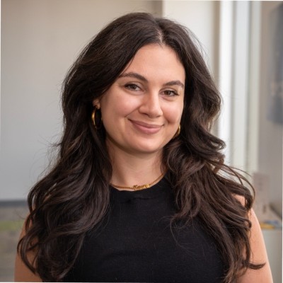 This is a headshot of Dr. Laura Baehr. She is a white woman with long dark hair and brown eyes. She is smiling and wearing a black top, gold hoop earrings, and a gold chain necklace.