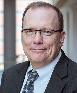 Portrait of Bill Cooper wearing a dark suit jacket, a white dress shirt, and a patterned tie, standing indoors with a blurred hallway background