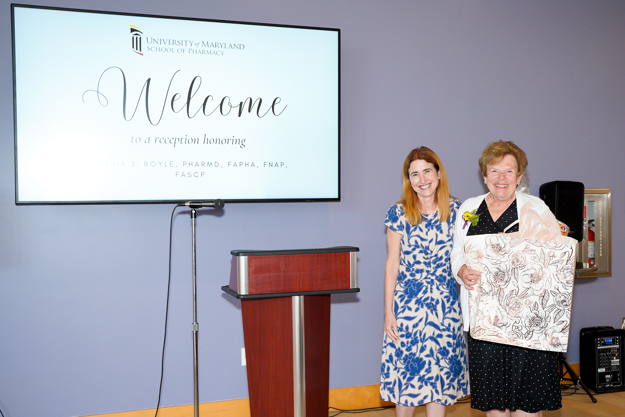 Dean Sarah L.J. Michel and Cynthia Boyle standing at a reception in Pharmacy Hall in front of a projector screen that says 
