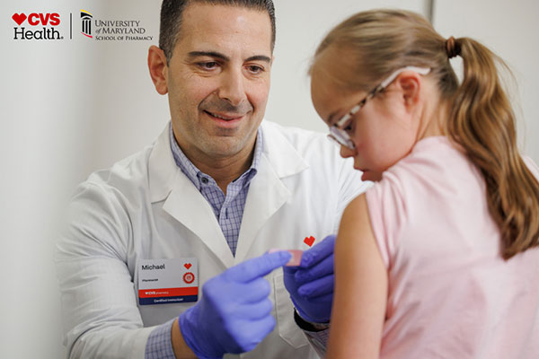 Pharmacist placing a bandage on a patient’s arm with CVS Health and UMD School of Pharmacy logos.