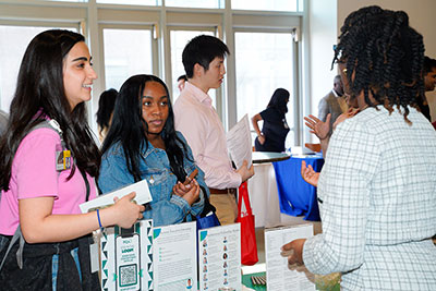 Student pharmacists speak with a recruiter at a career fair table, holding resumes and informational materials in a bright indoor event space