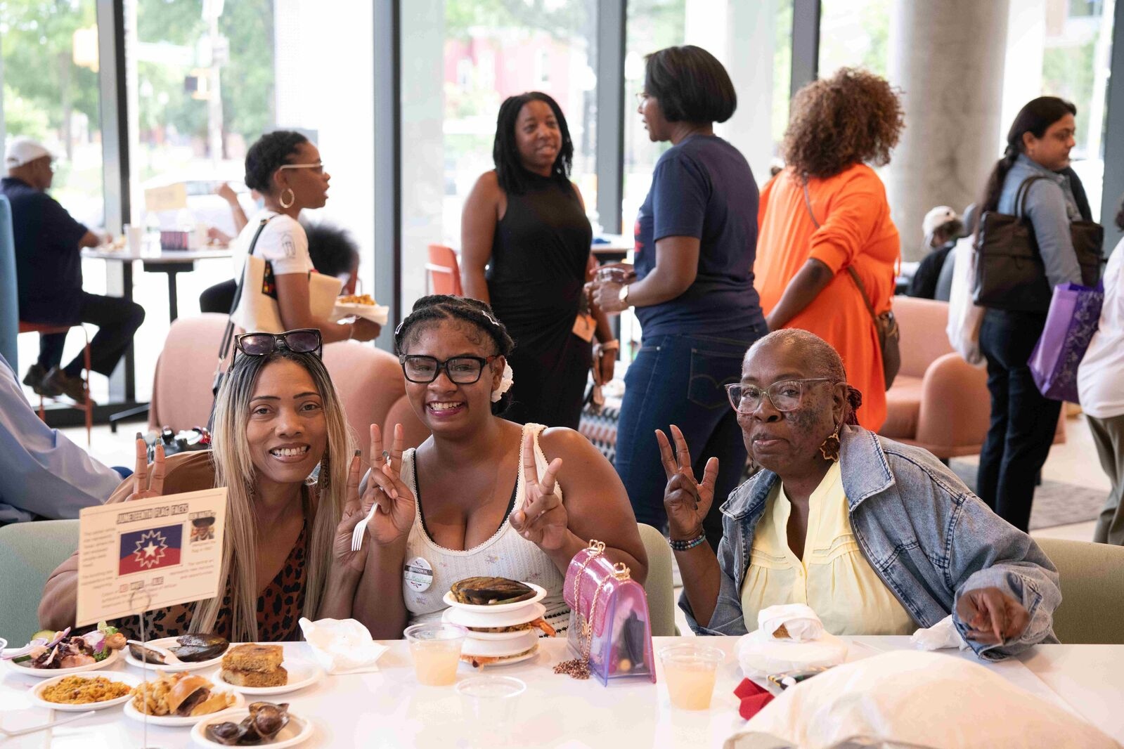 three women smiling giving peace signs with their hands at a dinner table in a brightly lit event space.