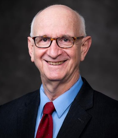 Jonathan Garlick, a man wearing dark full-rim glasses, a dark suit, blue shirt, and red tie, sits for a headshot photo