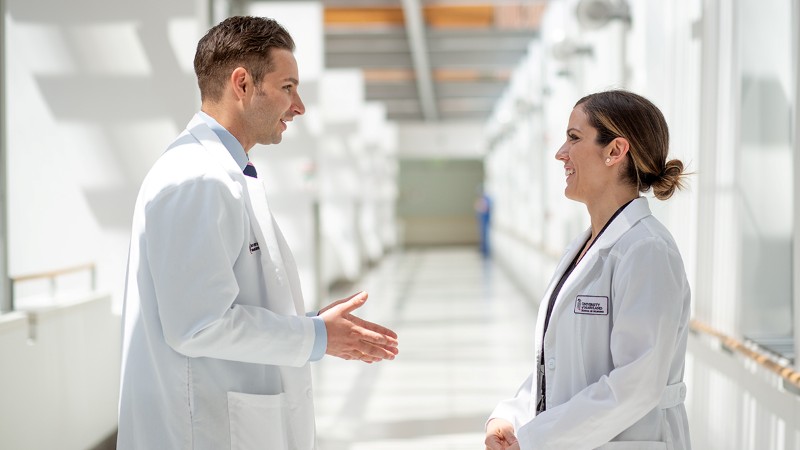 Two smiling nurse practitioners in white coats converse in a bright hospital corridor, conveying teamwork, professionalism, and friendly collaboration.