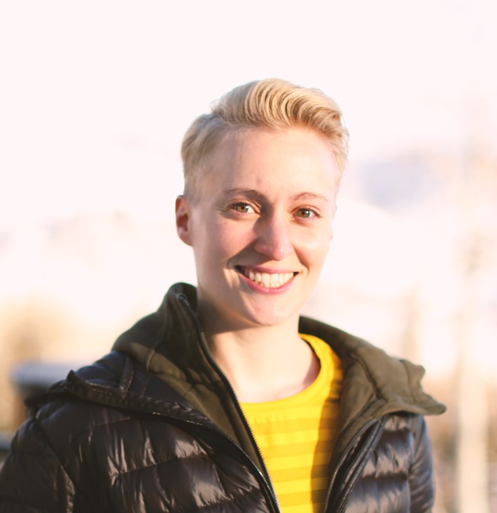 Paige Jarreau smiles into the camera, with short, light-blonde hair, standing outdoors in sunlight and wearing a black puffer jacket over a bright yellow top, in front of a softly blurred background