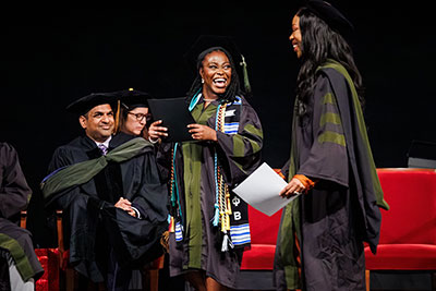 Sandeep Devabhakthuni sits at left on stage as a graduate receives recognition during a PharmD graduation ceremony.