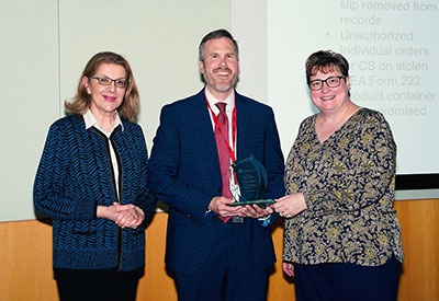 Fadia Shaya, Justin Wood of the DEA, and Jill Morgan stand together as Wood holds an award following his presentation.