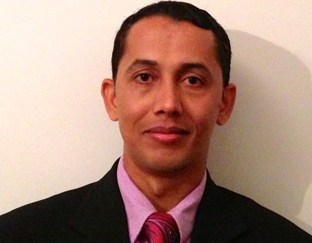 Professional photograph of a Dr. Shyam Sharma wearing a black suit jacket, a light purple dress shirt, and a red patterned tie against a plain white background.