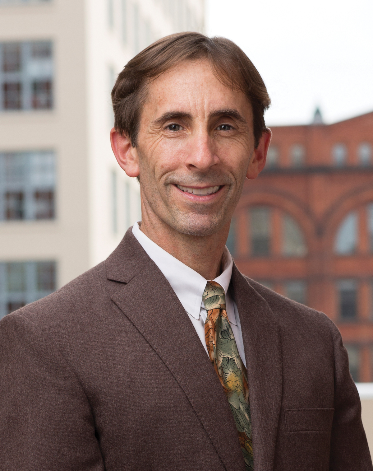 Daniel Mullins, PhD, wearing a brown suit, standing outside with city buildings in the background