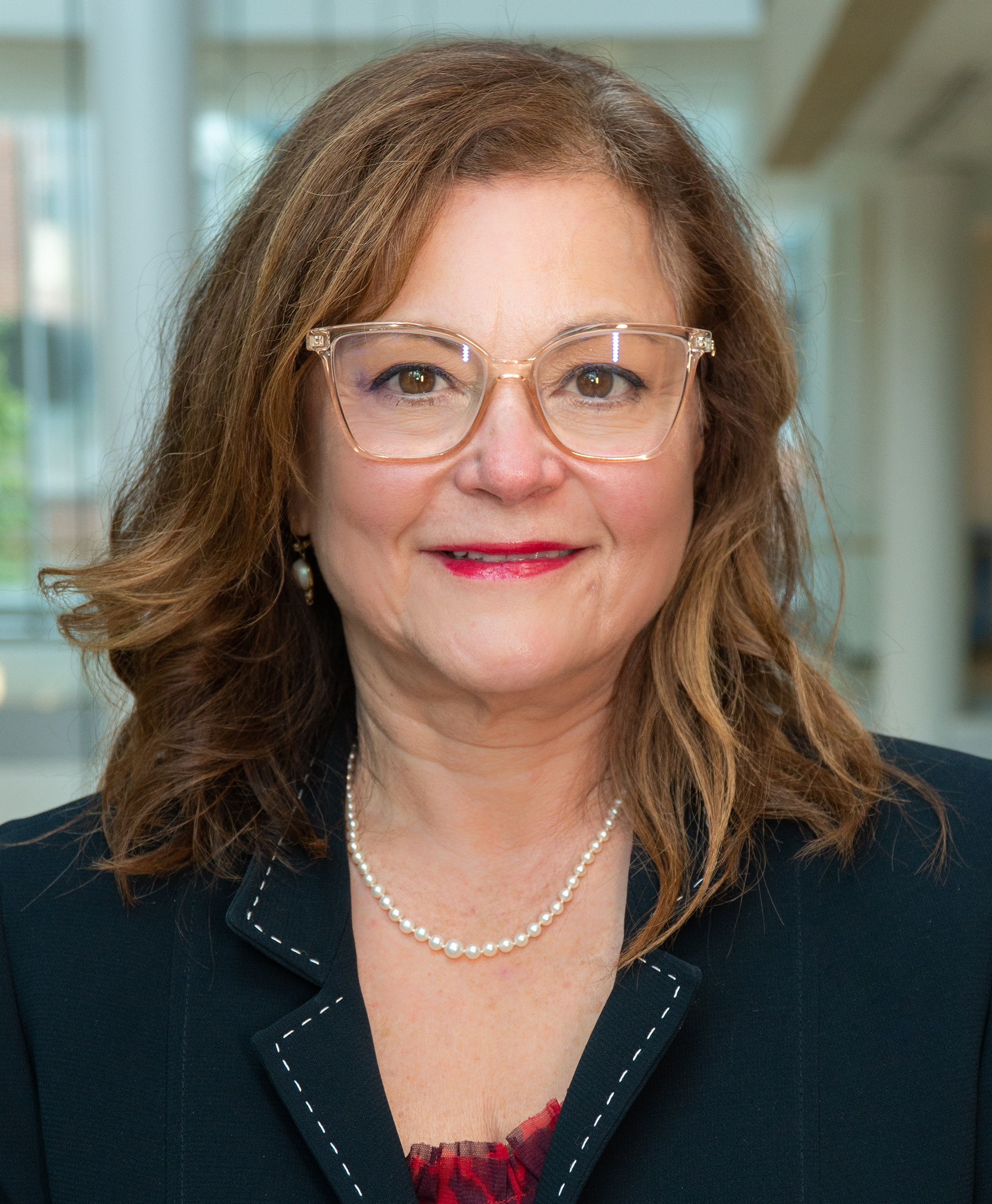 Magaly Rodriguez de Bittner, wearing a dark blazer and light glasses, posed inside with a blurred background of windows.