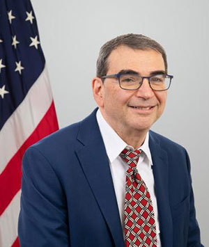 Steven Kozlowski wearing a navy blue jacket and glasses, with a U.S. flag in the background
