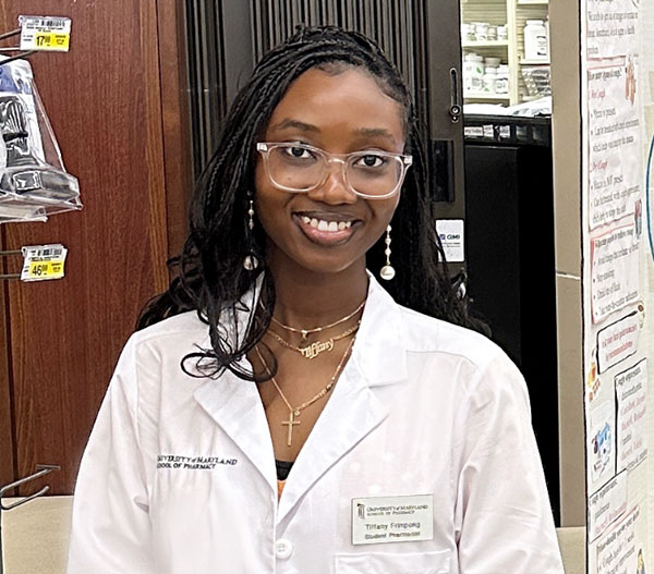 Tiffany Frimpong in a white pharmacy lab coat standing in a pharmacy workspace with shelves of medications in the background.