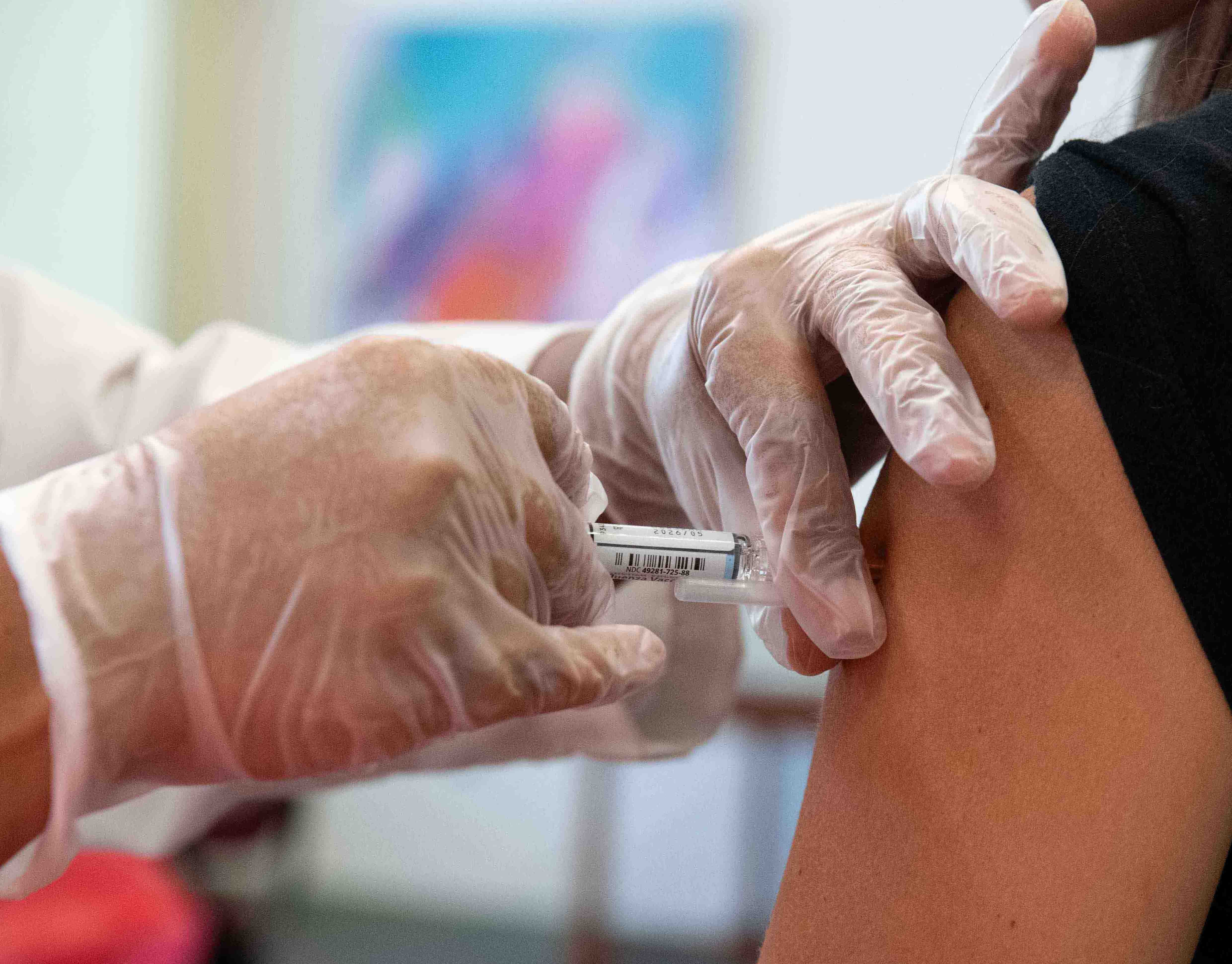 Photograph showing a healthcare professional wearing gloves administering a vaccine injection into a person's upper arm.