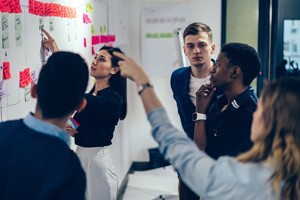 A focused group of young professionals collaborates, examining a whiteboard covered with colorful sticky notes and diagrams in a modern office setting.