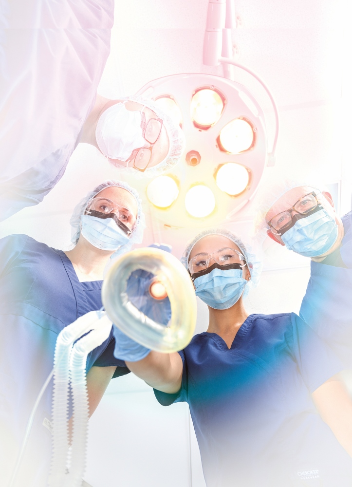 Four masked and gowned nurse anesthetists, viewed from below, gather under a bright surgical light, conveying focus and teamwork in an operating room.