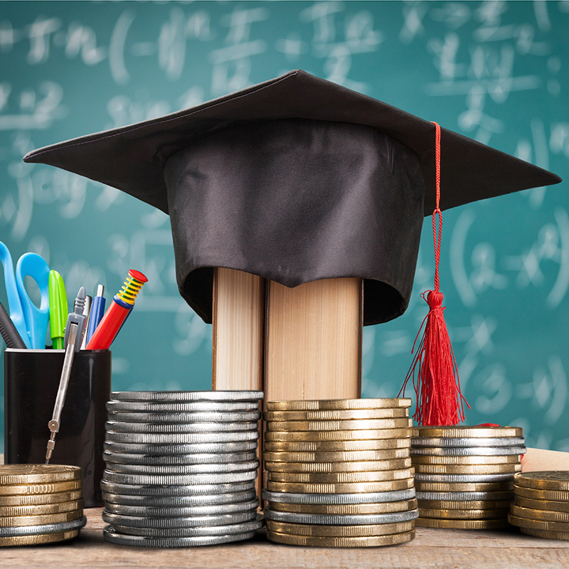 A graduation cap on two books sits behind stacks of coins, with a cup of school supplies and a chalkboard in the background, symbolizing education costs or scholarships.