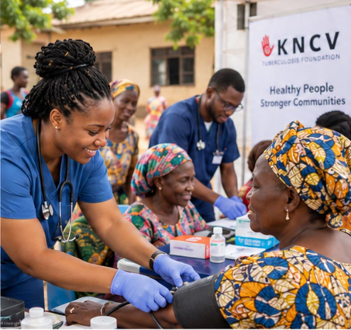 At a community health outreach, a smiling nurse in blue checks a seated woman’s blood pressure as clinicians assist nearby; a KNCV banner stands behind them.