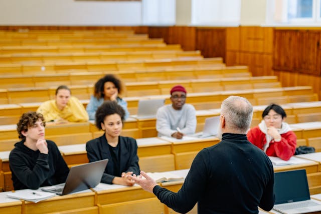 A man stands at the front of a room lecturing while an audience of people sit and listen attentively to him.