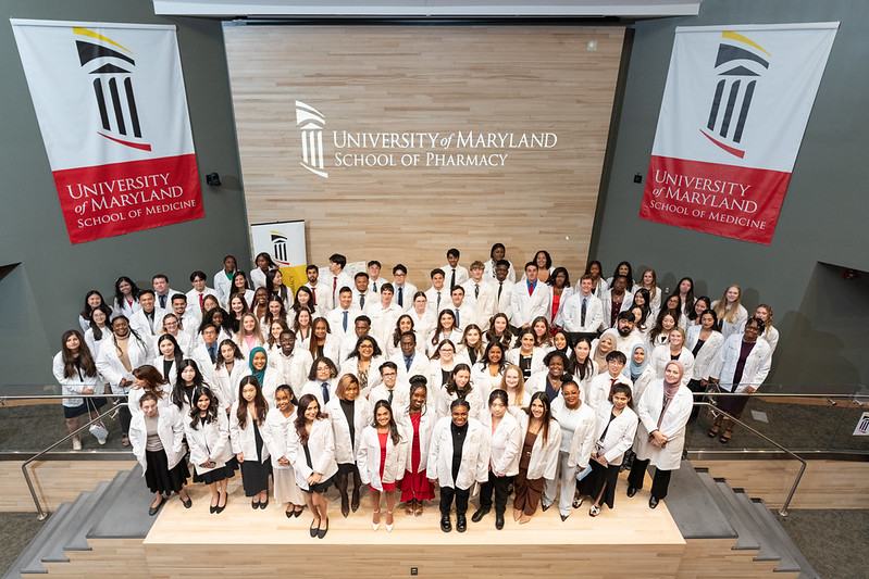 A large group of student pharmacists wearing white coats stand on a stage facing the camera.