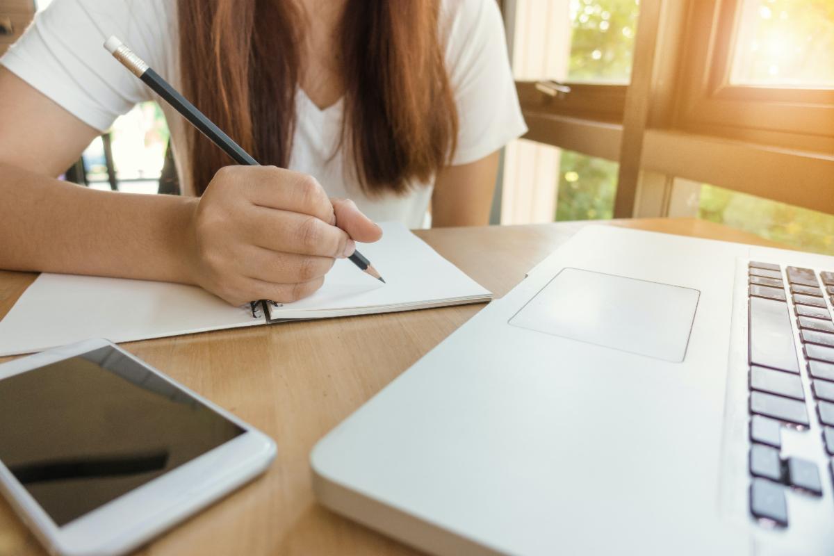 Individual has a pencil in hand posed above a notebook while facing a laptop.