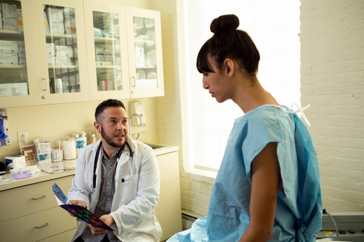 A transmasculine doctor pointing to medical documents while speaking to a transfeminine patient in a medical gown.