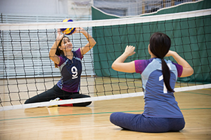 Two women sitting on a gym floor with a new between them as one of the women hits a volleyball