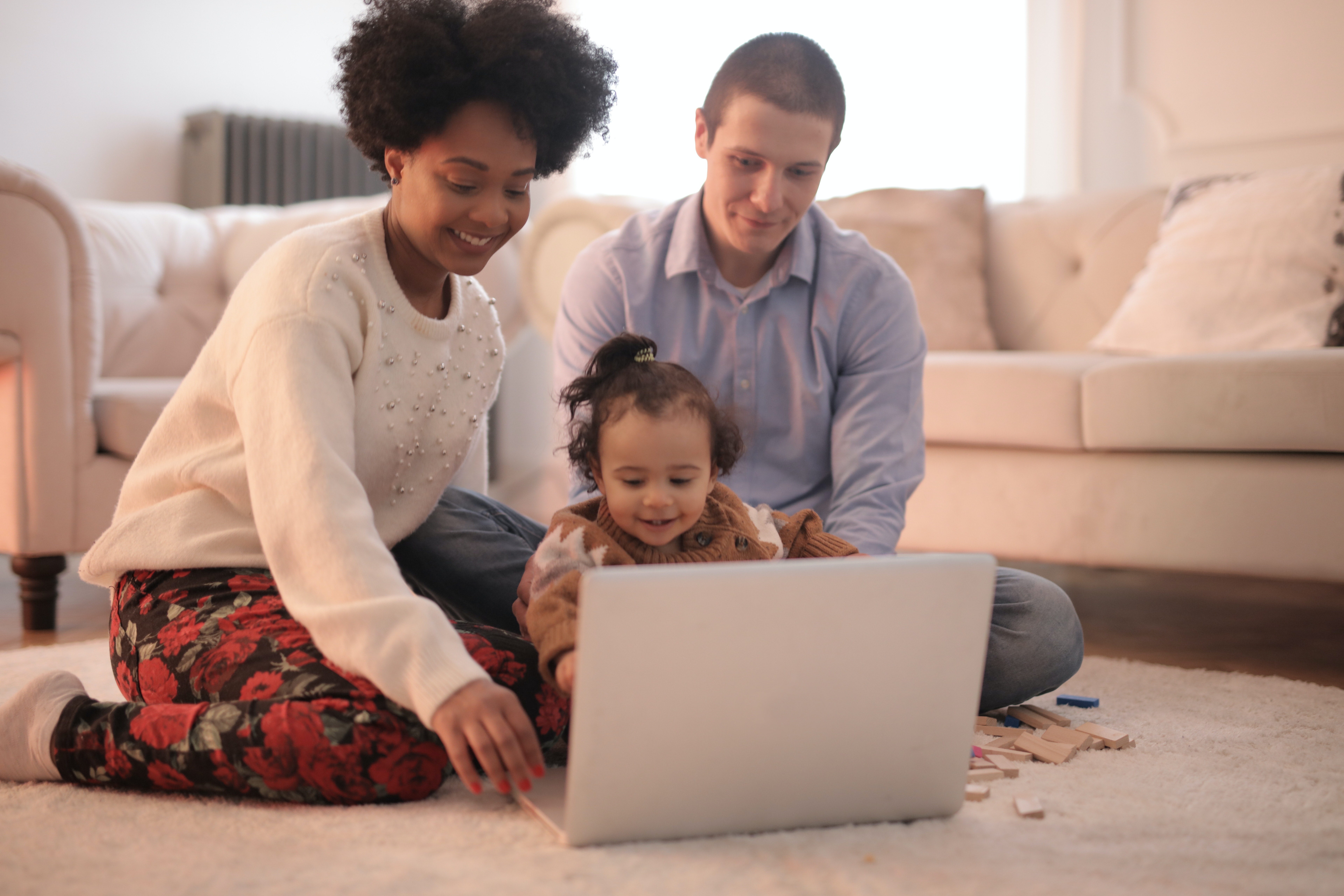 Two parents sit on the floor with a baby. The mother points and smiles at a laptop that is on the floor.