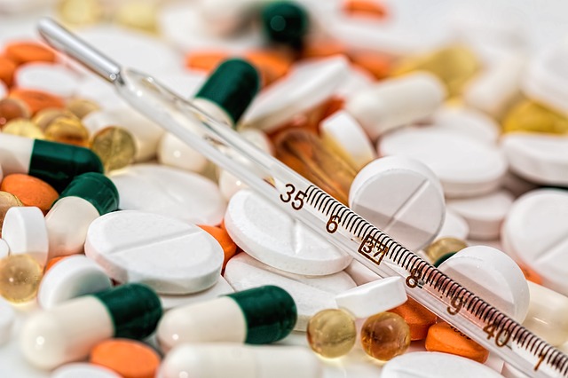 Multi-colored tablets and capsules spilled out across a flat surface with a glass thermometer placed on top of them.