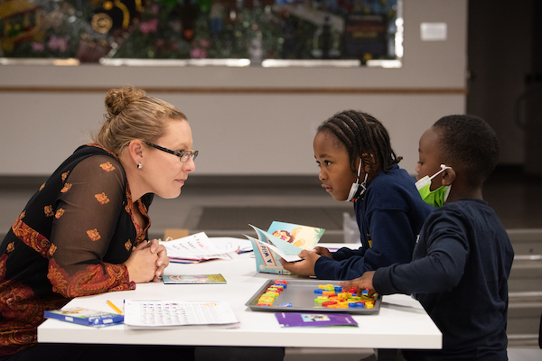 white woman with brown hair in brown dress with orange accents, tutoring black child with braids