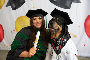 Caroline Benzel in School of Medicine regalia holding a rolled up piece of paper with her Rottweiler Loki, wearing a doctor's coat and mortarboard