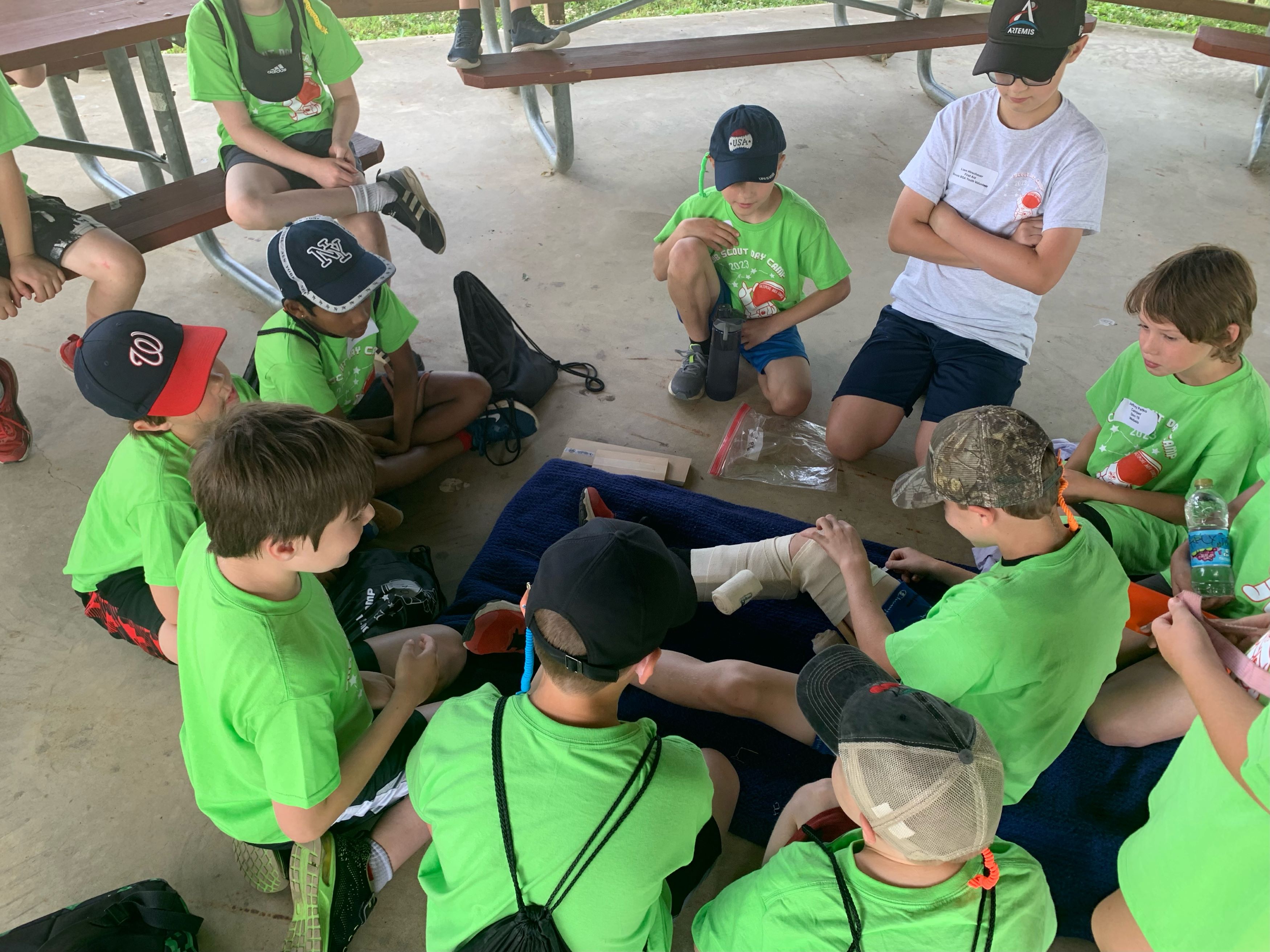 Scouts in green shirts gathered around a mat on which a scout is bandaging his leg