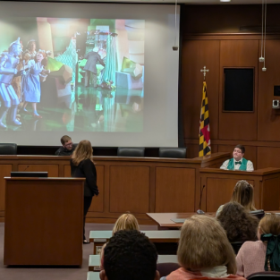 Man dressed as Wizard of Oz being questioned in courtroom in front of lawyer, judge, with photo from 1939 Oz movie shown as evidence
