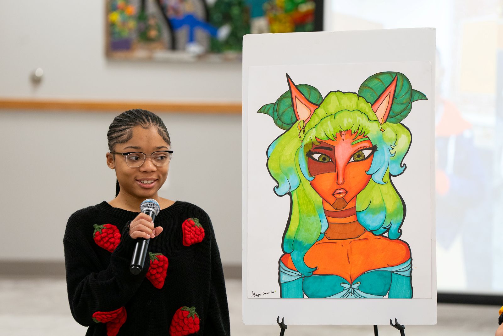 A young girl wearing glasses and a strawberry sweater is holding a microphone as she presents her artwork. The piece is a stylized portrait of a woman with bright green hair and horns.