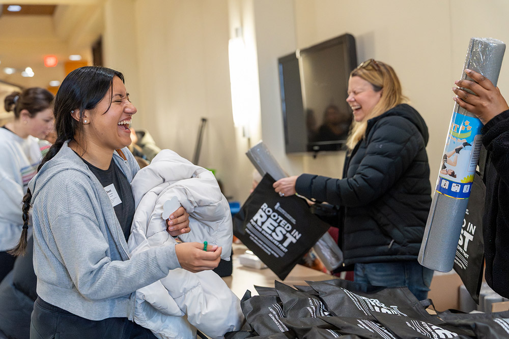 A woman laughs and holds a white jacket while interacting with others at a lively event. They hold yoga mats and tote bags, conveying a joyful atmosphere.