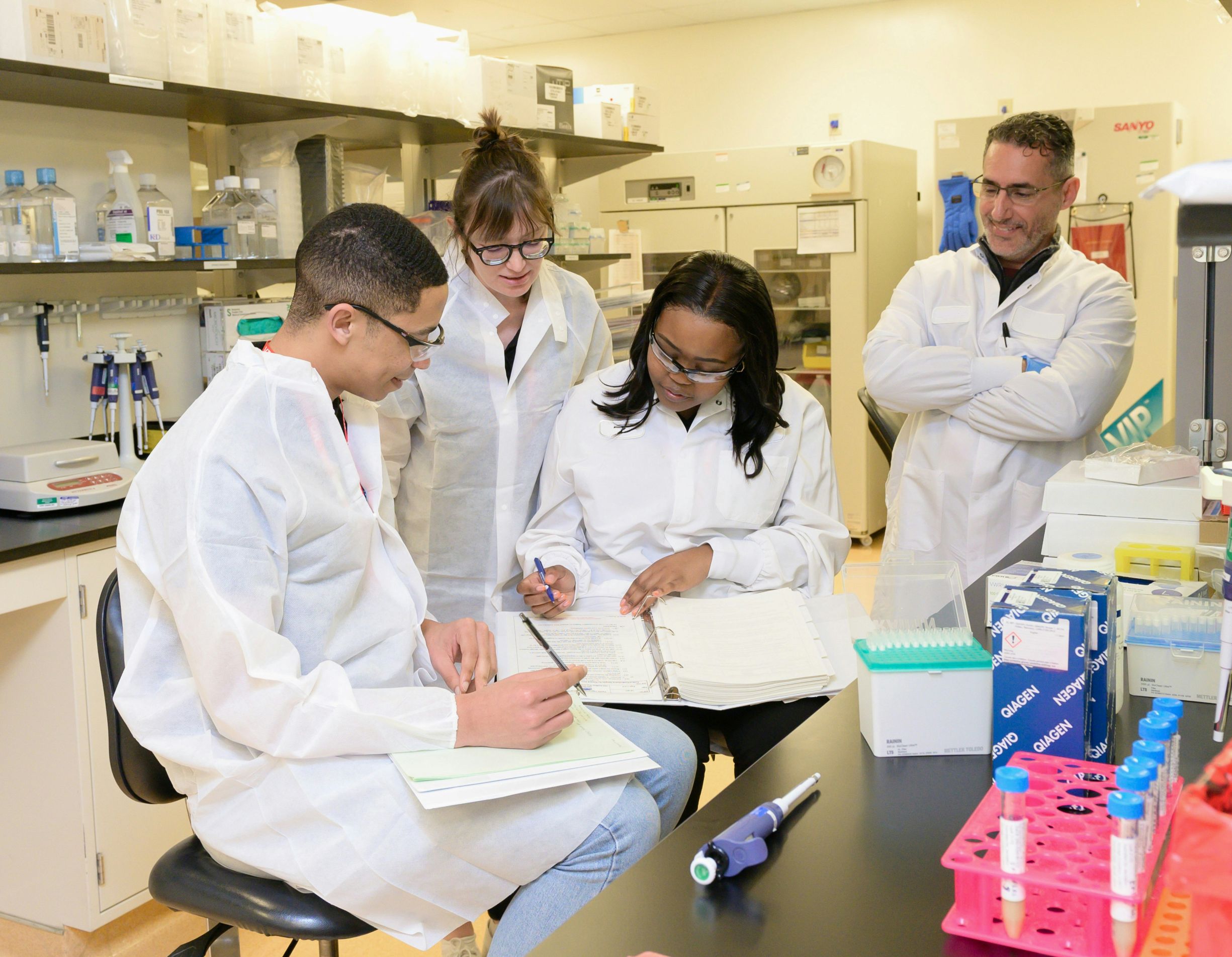 Four scientists in lab coats review notes together in a laboratory, surrounded by research equipment, indicating collaborative scientific work